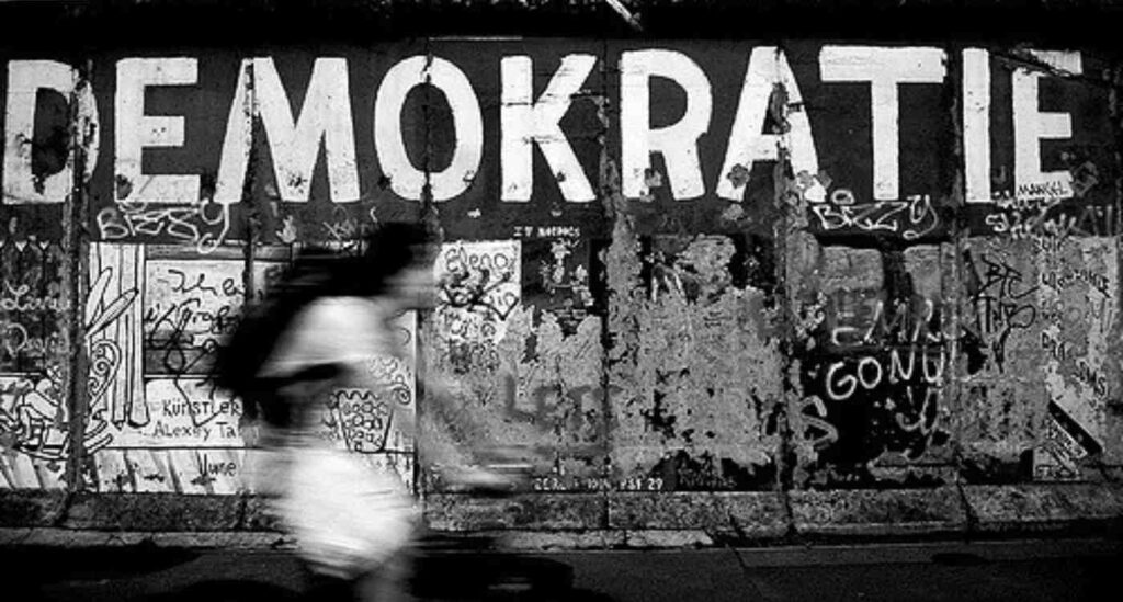 A cyclist passing under a graffito of the German word "Demokratie"