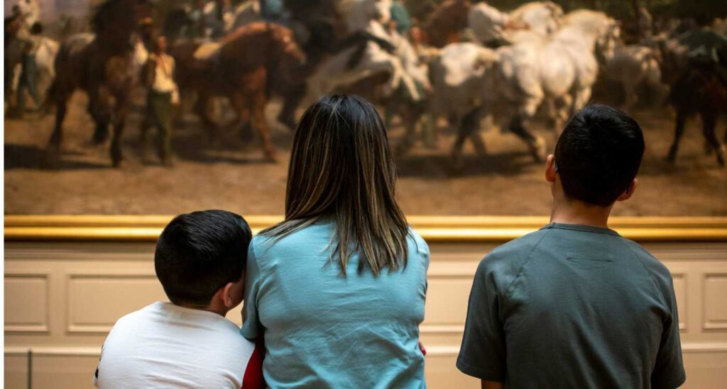 Image of an adult and two children looking at a painting featuring horses.