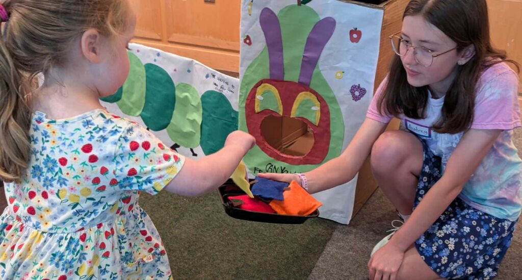 A teen summer volunteer hands a young child bean bags to play a Very Hungry Caterpillar tossing game at the 2025 TSV Eric Carle Party event.