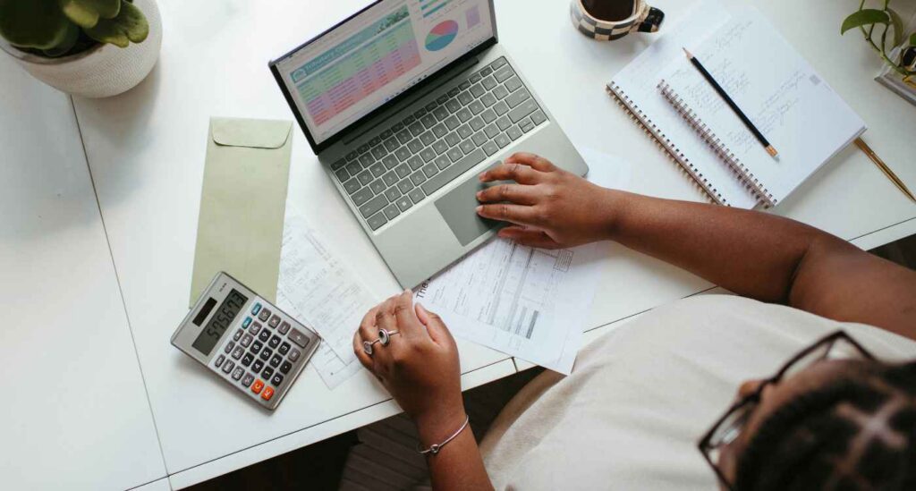 Person looking at a spreadsheet on a laptop screen with notebooks and a calculator on the table beside them.